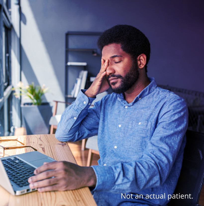 Man sitting at a desk with a laptop, rubbing his eyes and appearing fatigued or uncomfortable, possibly experiencing eye strain or dry eye symptoms. Caption reads "Not an actual patient."